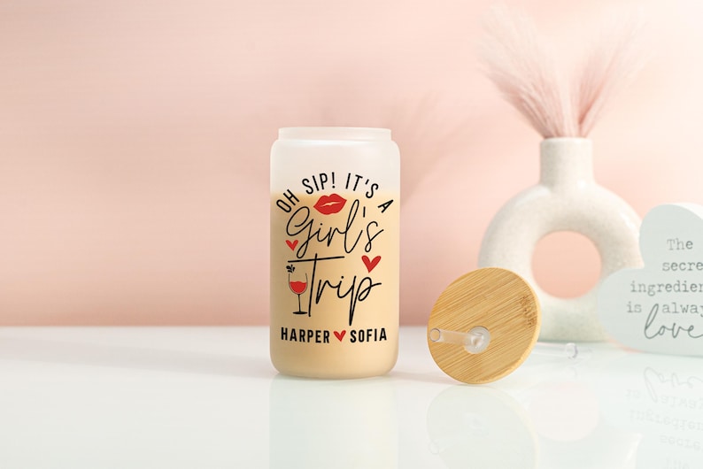 May include: A frosted glass can-shaped tumbler with a bamboo lid. The tumbler features the words "OH SIP! IT'S A Girl's Trip" in black and red script, with a red lip graphic and a wine glass illustration. The names "Harper" and "Sofia" are below.