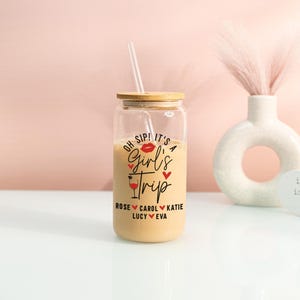 May include: A glass tumbler with a bamboo lid and straw, containing a light-coloured drink. The tumbler is printed with "Oh Sip! It's a Girl's Trip" and names. A vase and heart-shaped sign are in the background.