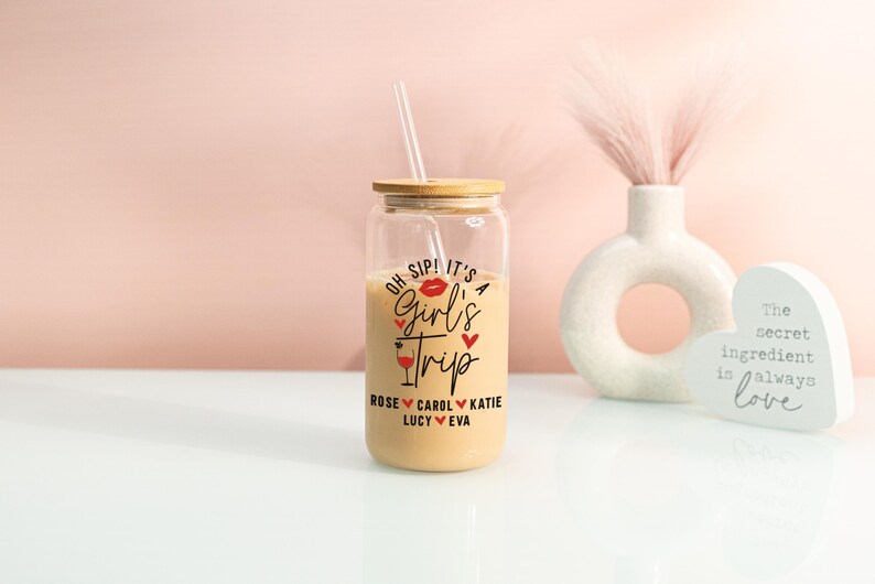 May include: A glass tumbler with a bamboo lid and straw, containing a light-coloured drink. The tumbler is printed with "Oh Sip! It's a Girl's Trip" and names. A vase and heart-shaped sign are in the background.