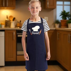 May include: A navy blue apron with a white chef's hat, fork, and rolling pin design. The name "Daniel" is printed below the chef's hat. The apron is worn over a striped shirt and jeans.