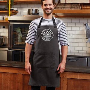 Op de afbeelding: Een donkergrijze schort met een grote voorzak en zwarte bandjes. De schort heeft een wit cirkelvormig logo met de tekst "GEORGE Steak House" en gekruiste vleesmessen. De persoon draagt een gestreept shirt.