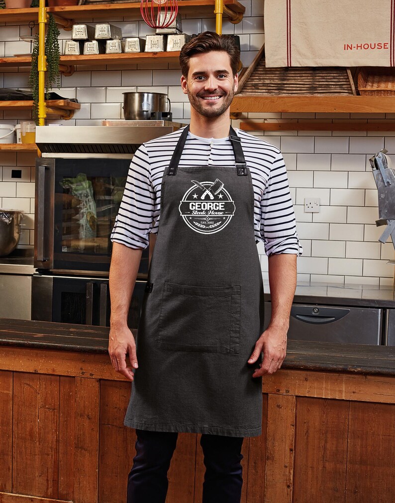 Op de afbeelding: Een donkergrijze schort met een grote voorzak en zwarte bandjes. De schort heeft een wit cirkelvormig logo met de tekst "GEORGE Steak House" en gekruiste vleesmessen. De persoon draagt een gestreept shirt.