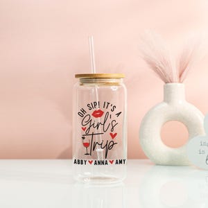 May include: A clear glass tumbler with a bamboo lid and a straw. The tumbler features the text "Oh Sip! It's a Girl's Trip" in black and red, with the names Abby, Anna, and Amy below. A decorative vase and heart-shaped sign are in the background.