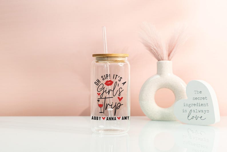 May include: A clear glass tumbler with a bamboo lid and a straw. The tumbler features the text "Oh Sip! It's a Girl's Trip" in black and red, with the names Abby, Anna, and Amy below. A decorative vase and heart-shaped sign are in the background.