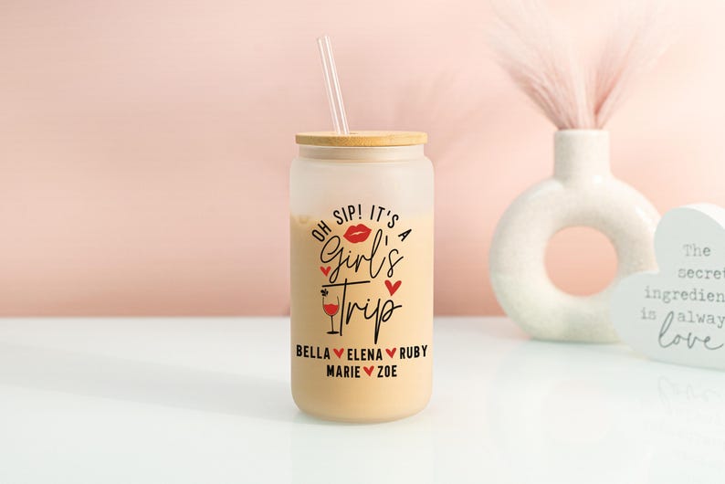 May include: A frosted glass can-shaped tumbler with a bamboo lid and a clear straw. The tumbler is filled with a light-coloured beverage and features the text "Oh Sip! It's a Girl's Trip" in black and red, with friends' names.