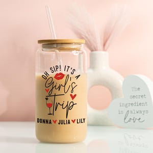 May include: A clear glass tumbler with a wooden lid and a straw, filled with a creamy drink. The tumbler has the text "OH SIP! IT'S A Girl's Trip" with names below. A decorative vase and heart-shaped sign are in the background.