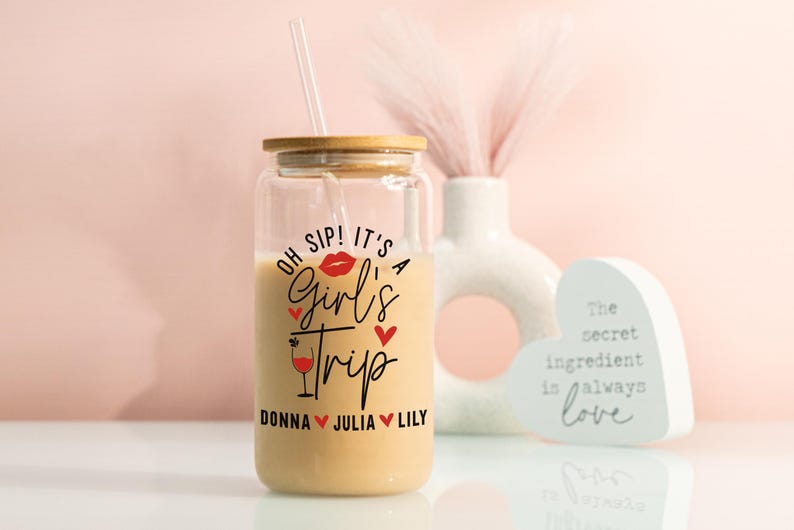 May include: A clear glass tumbler with a wooden lid and a straw, filled with a creamy drink. The tumbler has the text "OH SIP! IT'S A Girl's Trip" with names below. A decorative vase and heart-shaped sign are in the background.