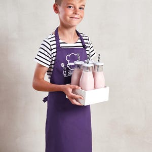 May include: A purple apron with a chef logo and text is worn by a person holding a white tray with glass bottles of pink liquid. The bottles have white lids and black and white striped straws. The person is wearing a black and white striped shirt.