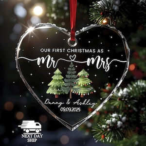 May include: Heart-shaped clear glass Christmas ornament with the text "OUR FIRST CHRISTMAS AS Mr & Mrs" and the names "Danny & Ashley" with the date "09.09.2025". The ornament features three Christmas trees and a red ribbon for hanging.