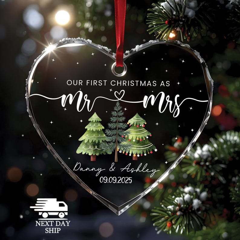 May include: Heart-shaped clear glass Christmas ornament with the text "OUR FIRST CHRISTMAS AS Mr & Mrs" and the names "Danny & Ashley" with the date "09.09.2025". The ornament features three Christmas trees and a red ribbon for hanging.