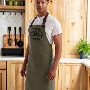 Op de afbeelding: Een olijfgroene schort met een bruine leren nekriem en een zak. De schort heeft een cirkelvormig logo met de tekst "HENRY Steak House" en gekruiste gereedschappen. De persoon draagt een wit shirt en een blauwe jeans.