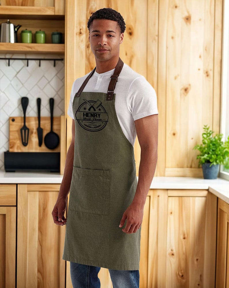 Op de afbeelding: Een olijfgroene schort met een bruine leren nekriem en een zak. De schort heeft een cirkelvormig logo met de tekst "HENRY Steak House" en gekruiste gereedschappen. De persoon draagt een wit shirt en een blauwe jeans.