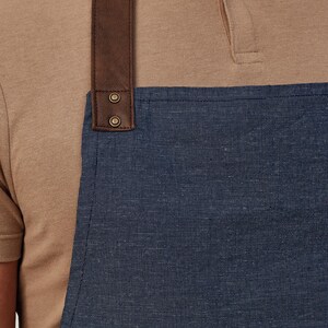 Op de afbeelding: Close-up van een blauwe denim schort met een bruine leren band. De band is met twee metalen klinknagels aan de schort bevestigd. De schort wordt gedragen over een bruin T-shirt met korte mouwen.
