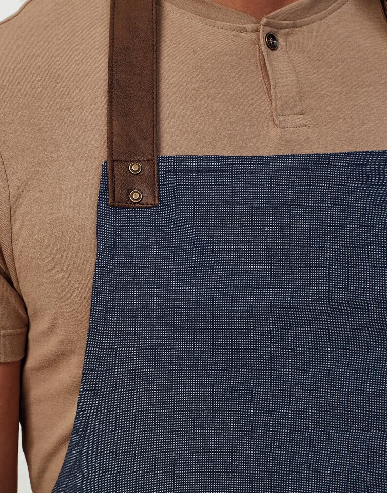 Op de afbeelding: Close-up van een blauwe denim schort met een bruine leren band. De band is met twee metalen klinknagels aan de schort bevestigd. De schort wordt gedragen over een bruin T-shirt met korte mouwen.