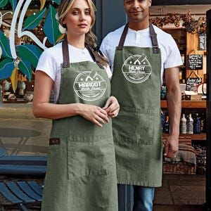 Op de afbeelding: Twee mensen dragen olijfgroene schorten met bruine leren bandjes. Elke schort heeft een grote voorzak en een cirkelvormig logo met tekst. De schorten worden gedragen over witte t-shirts. De logo's luiden "MARGOT Steak House" en "HENRY Steak House".
