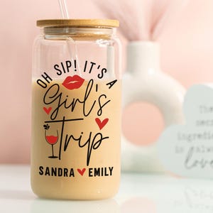 May include: A clear glass tumbler with a bamboo lid and straw, filled with a creamy drink. The tumbler has the text "Oh Sip! It's a Girl's Trip" with red heart and lip graphics, and personalized names. A fun gift idea for a friend.