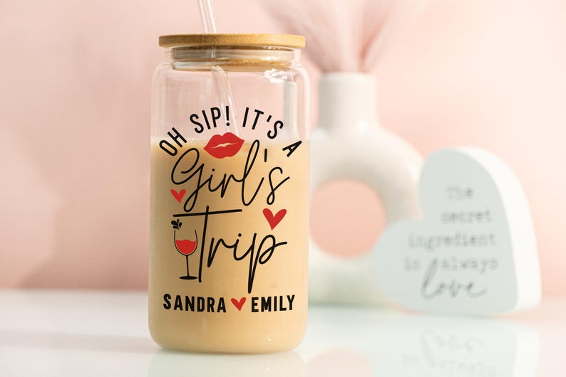 May include: A clear glass tumbler with a bamboo lid and straw, filled with a creamy drink. The tumbler has the text "Oh Sip! It's a Girl's Trip" with red heart and lip graphics, and personalized names. A fun gift idea for a friend.