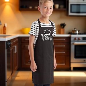 May include: A black apron with a chef graphic and the name "KEVIN" in white. The apron is worn over a striped shirt and jeans. The background shows a kitchen with wooden cabinets and stainless steel appliances.