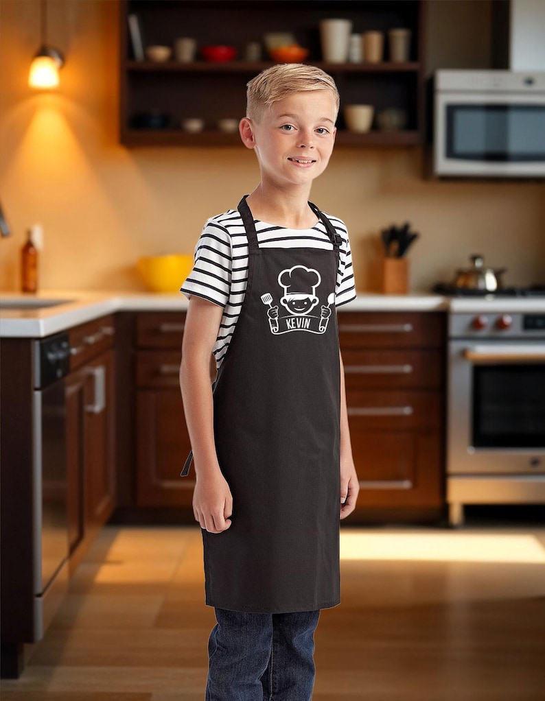 May include: A black apron with a chef graphic and the name "KEVIN" in white. The apron is worn over a striped shirt and jeans. The background shows a kitchen with wooden cabinets and stainless steel appliances.