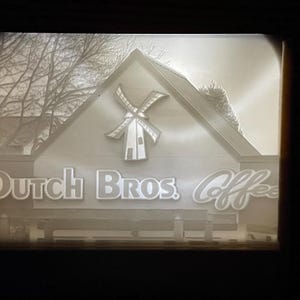 May include: A grayscale image of a Dutch Bros. Coffee shop. The shop features a windmill logo and the words "Dutch Bros. Coffee" in a stylized font. A bench is visible in front of the shop, and bare tree branches are in the background.