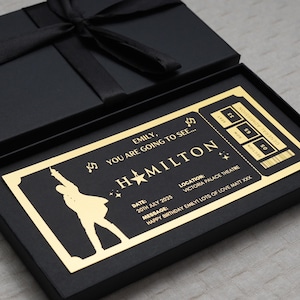 May include: A gold-coloured ticket for a Hamilton musical performance. The ticket is in a black box with a black ribbon. The ticket says "Emily, you are going to see... Hamilton" and includes the date, location, and a message.