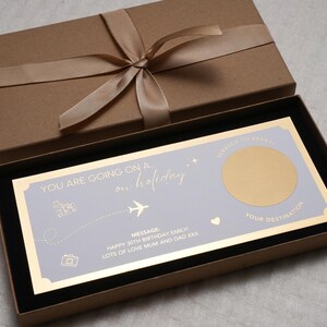 May include: A light blue and gold card with the text "YOU ARE GOING ON A on holiday". The card has a gold circle with a scratch-off surface that says "SCRATCH TO REVEAL YOUR DESTINATION". The card is in a brown box with a gold ribbon.