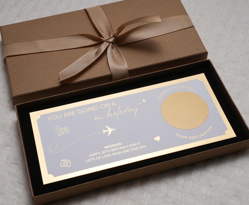 May include: A light blue and gold card with the text "YOU ARE GOING ON A on holiday". The card has a gold circle with a scratch-off surface that says "SCRATCH TO REVEAL YOUR DESTINATION". The card is in a brown box with a gold ribbon.