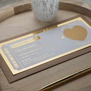 Op de afbeelding: Een gouden boarding pass met een grijze achtergrond. De tekst op de boarding pass luidt "Boarding Pass", "You are going to", "Passenger Name: Emily", "Duration: 5 Nights", "Message: Happy 30th Birthday Emily, Lots of Love Mum and Dad xxx". Er is een gouden hart dat je kunt schrapen om een verborgen bericht te onthullen.