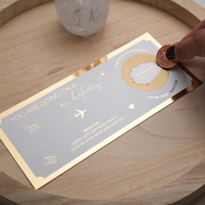 May include: A gold and gray scratch-off card with the text "You are going on a holiday" and "Scratch to reveal your destination". The card has a gold coin on top of the scratch-off area.