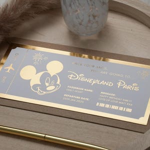 May include: A gold-trimmed invitation card with a Mickey Mouse illustration and the text "Pack your bags, you are going to Disneyland Paris". The card also includes the text "Passenger Name: Emily Heart" and "Departure Date: 25th Jan 2025".