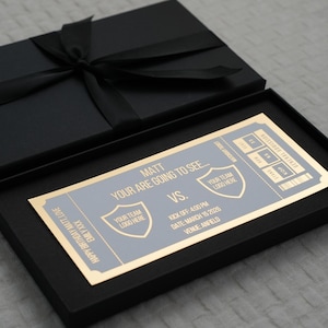 Op de afbeelding: Een zwarte geschenkdoos met bijpassend deksel en zwart lint. Binnenin staat een ticket met gouden rand en grijze tekst met de tekst "Matt, Your are going to see..." met teamlogo's en evenementdetails.