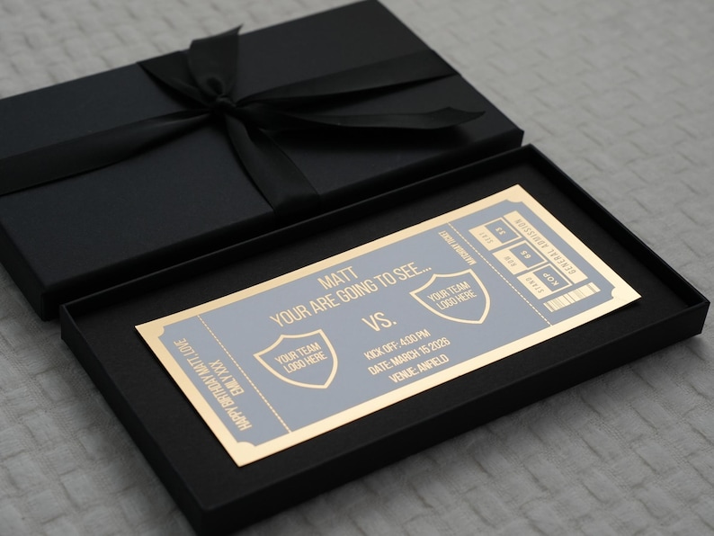 Op de afbeelding: Een zwarte geschenkdoos met bijpassend deksel en zwart lint. Binnenin staat een ticket met gouden rand en grijze tekst met de tekst "Matt, Your are going to see..." met teamlogo's en evenementdetails.