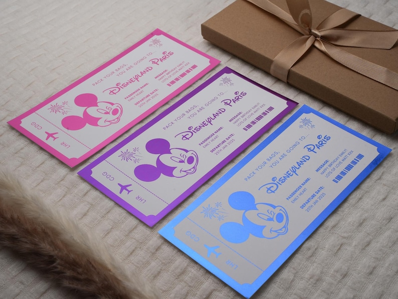 May include: Three colorful paper airplane tickets with Mickey Mouse on each. The tickets are pink, purple, and blue. The text on the tickets reads "Pack your bags, you are going to Disneyland Paris".