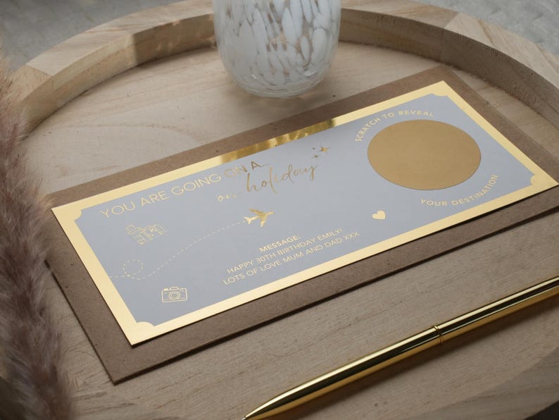 May include: A light blue and gold card with the text "YOU ARE GOING ON A HOLIDAY" and a gold circle with a scratch-off surface to reveal a hidden message. The card also includes a message that says "HAPPY 30TH BIRTHDAY EMILY LOTS OF LOVE MUM AND DAD XXX".