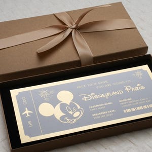 May include: A gold-colored ticket with a Mickey Mouse silhouette and the text "Pack your bags, you are going to Disneyland Paris". The ticket has a departure date of January 25th, 2025. The ticket is in a brown box with a gold ribbon.