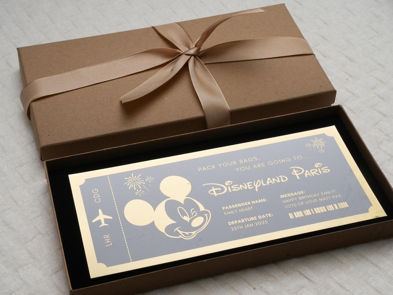May include: A gold-colored ticket with a Mickey Mouse silhouette and the text "Pack your bags, you are going to Disneyland Paris". The ticket has a departure date of January 25th, 2025. The ticket is in a brown box with a gold ribbon.