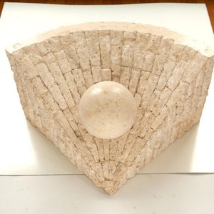 May include: A white stone sculpture of a sphere nestled in a fan-shaped, geometric pattern of stacked stone blocks.