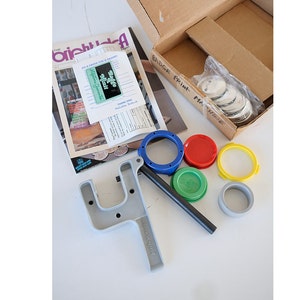 May include: A badge making kit with a gray plastic press, a blue plastic ring, a green plastic ring, a yellow plastic ring, a red plastic ring, and a box of metal badge blanks. The box has the text "Badge Print Machine" written on it.