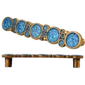 May include: A brass cabinet pull with a decorative design of blue circles. The pull has a curved shape and two mounting holes.