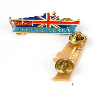 May include: Gold-colored pin featuring a ship with a Union Jack flag and the text "Reebok Fitness Cruiser".