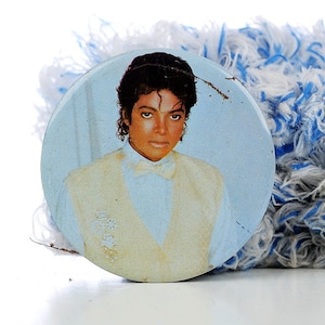 May include: A round, blue and white button with a picture of a man with dark hair wearing a white shirt and a light yellow vest. The button is slightly worn.