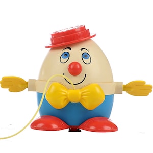 May include: A vintage pull toy shaped like an egg with a red hat, blue pants, yellow bow tie, and yellow hands. The toy has a smiling face with blue eyes and a red nose. The toy is attached to a yellow string.