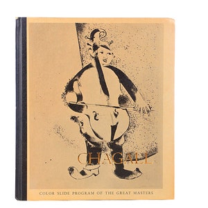 May include: A book with a tan cover featuring a black and white illustration of a figure playing a cello. The word "CHAGALL" is printed in gold, with "COLOR SLIDE PROGRAM OF THE GREAT MASTERS" below. The spine is black.