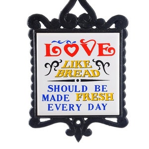 May include: Black cast iron sign with a white tile insert. The tile has a red, blue, and yellow graphic that says "Love like bread should be made fresh every day".