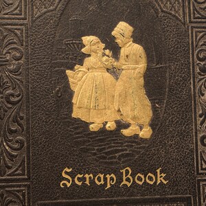 May include: A vintage leather-bound scrapbook with a gold embossed design featuring two figures in traditional Dutch clothing. The title "Scrap Book" is written in gold lettering on the front cover.