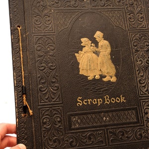 May include: A vintage black leather scrapbook with gold embossed details and a gold illustration of two people in traditional clothing. The cover reads "Scrap Book".