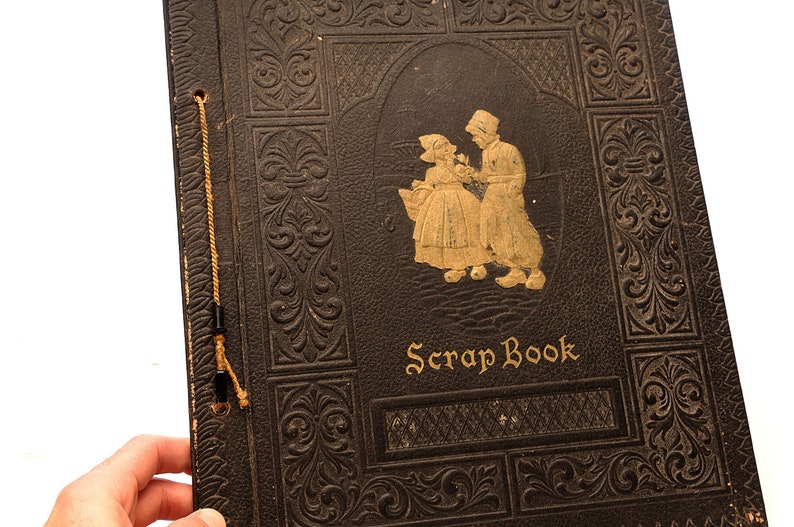 May include: A vintage black leather scrapbook with gold embossed details and a gold illustration of two people in traditional clothing. The cover reads "Scrap Book".