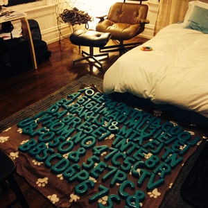 Peut inclure: Une couverture &agrave; motifs bruns et blancs avec des lettres en turquoise bleu qui &eacute;pellent les mots "PRESENT & MIX" et d'autres mots. La couverture est sur un sol en bois dans une pi&egrave;ce avec un lit blanc et un fauteuil en cuir brun.