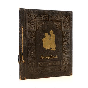 May include: A vintage brown leather-bound scrapbook with gold embossed details and a silhouette of two people on the front cover. The title "Scrap Book" is printed in gold lettering.