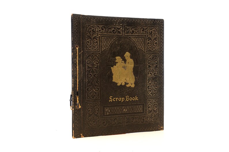 May include: A vintage brown leather-bound scrapbook with gold embossed details and a silhouette of two people on the front cover. The title "Scrap Book" is printed in gold lettering.